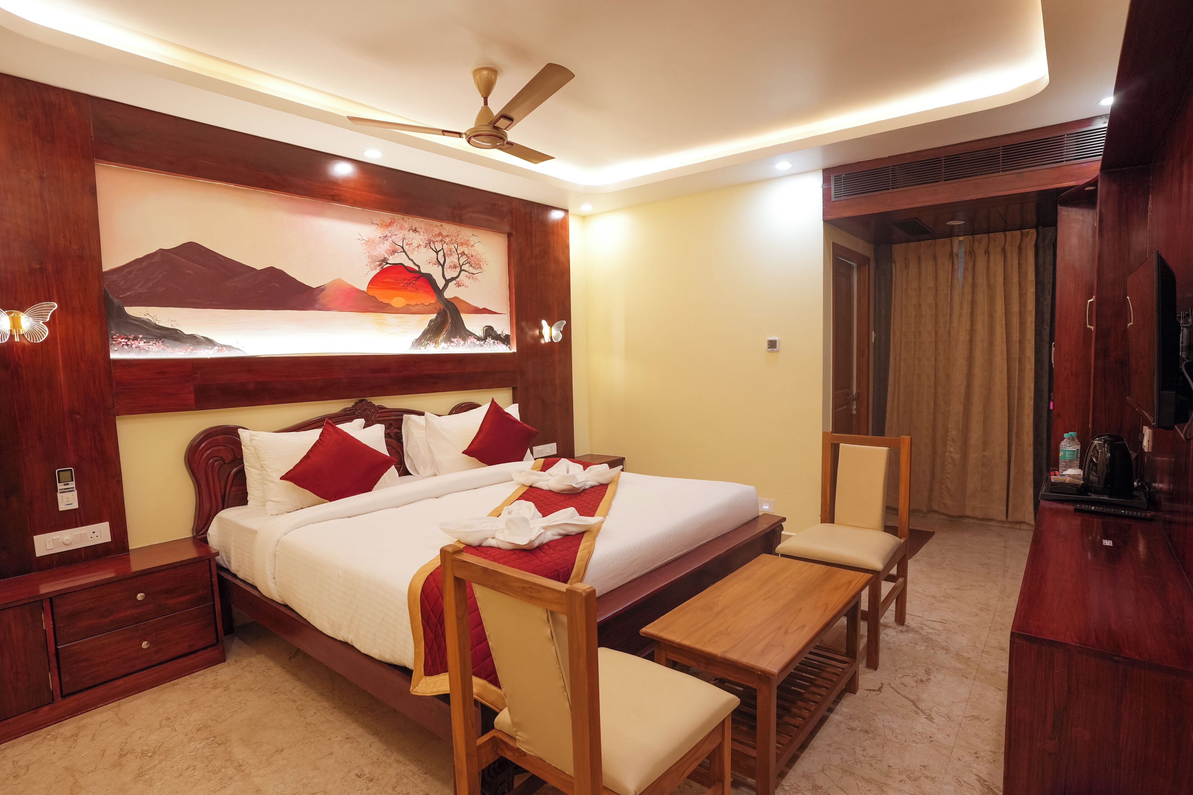 Habitación de hotel con cama, mesitas de noche, escritorio, silla, ventilador de techo y un cuadro en la pared.