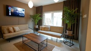 Een woonkamer met een bank, een salontafel, twee potplanten, een televisie waarop 'Rookie' te zien is, en een raam met gordijnen.