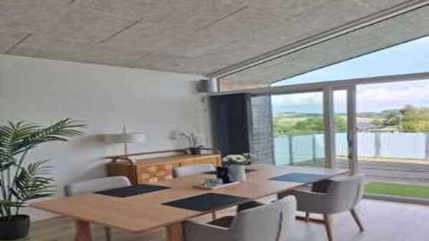 Un espace repas moderne avec une table et des chaises en bois, un petit coin cuisine doté d’un évier et de placards, et de grandes fenêtres offrant une vue sur l’extérieur.