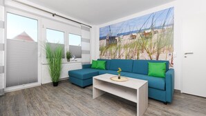 A modern living room with a blue sofa, a white coffee table, and a beach mural on the wall. - Moderne Ferienwohnung in Ruhiger Lage, Bensersiel (Esens)