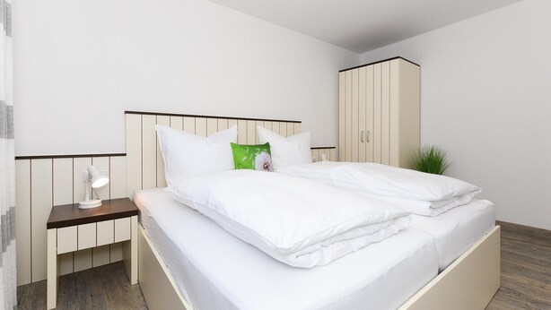 A neatly made bed with white bedding, a bedside table with a lamp, and a wardrobe in a modern, minimalistic room. - Moderne Ferienwohnung in Ruhiger Lage, Bensersiel (Esens)