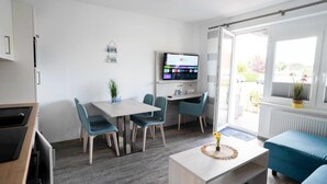A modern kitchen with a built-in oven, a dining area with a table and chairs, a TV mounted on the wall, and a large window providing a view of the outdoors. - Moderne Ferienwohnung in Ruhiger Lage, Bensersiel (Esens)
