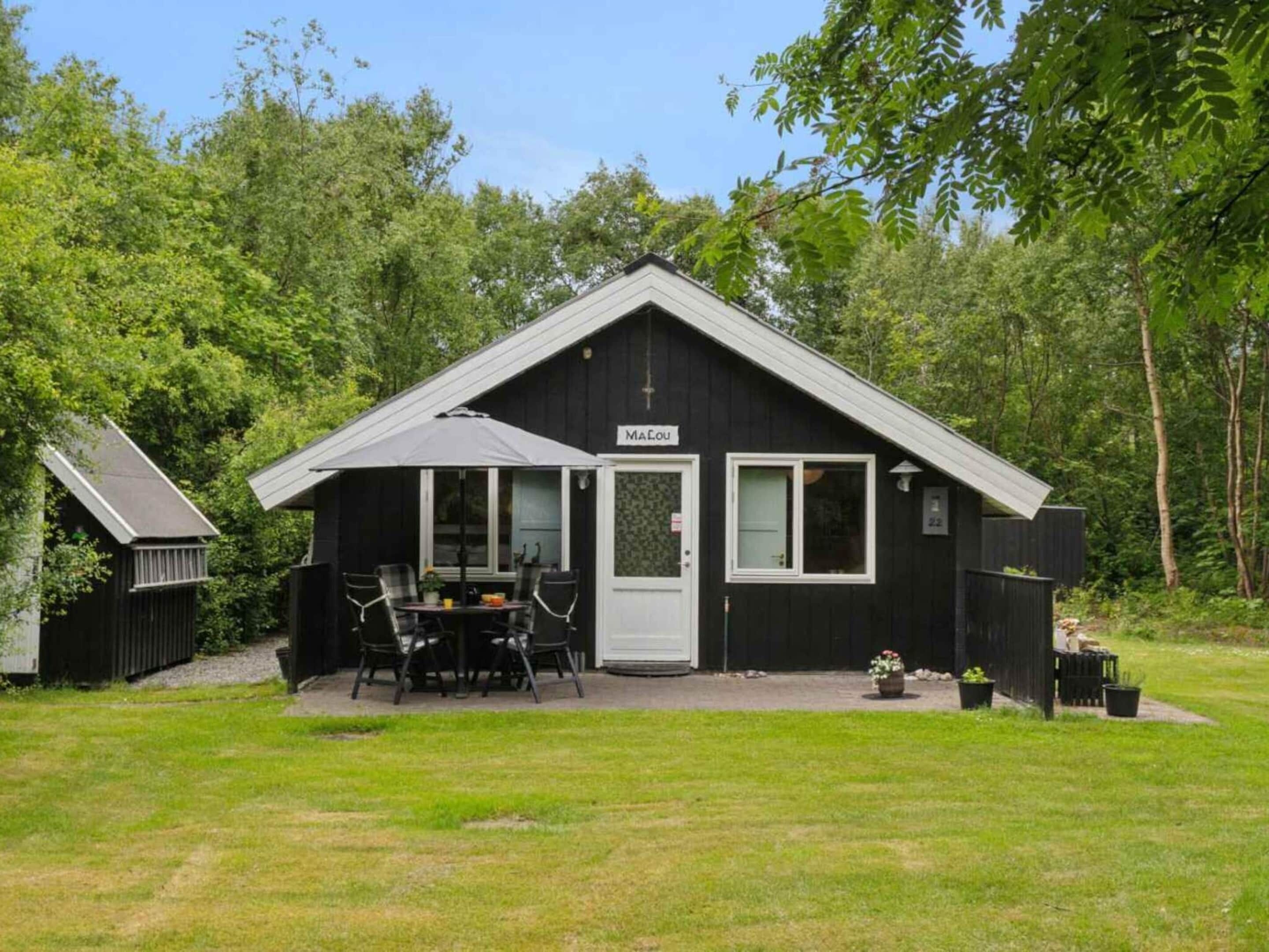 A small black building with a white door and windows, labeled "MaLou," with a patio table and chairs, surrounded by greenery.
