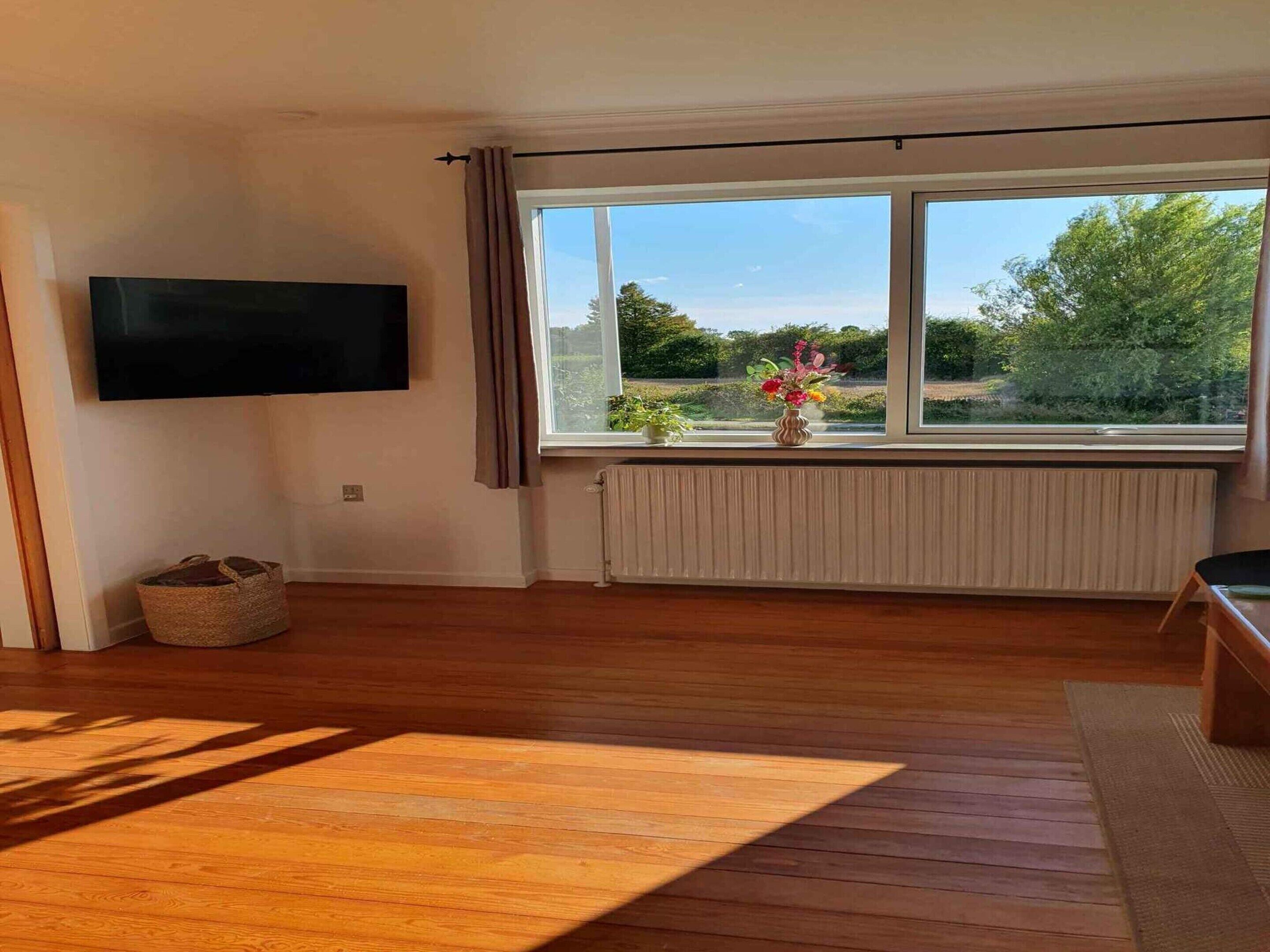 Ein geräumiges Wohnzimmer mit großem Fenster, einem wandmontierten Flachbildfernseher und Holzboden.