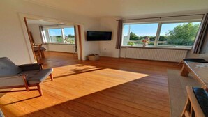 Un vaste salon doté d’un parquet, d’un téléviseur à écran plat fixé au mur et de grandes fenêtres donnant sur un paysage verdoyant.