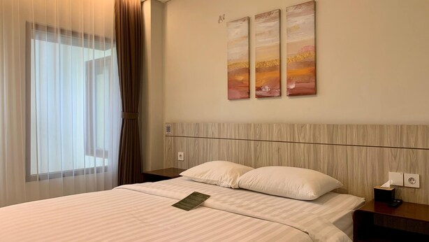A hotel room with a large bed, a nightstand, a wall-mounted power outlet, and a window with sheer curtains. - The Baliem Hotel (Jimbaran)