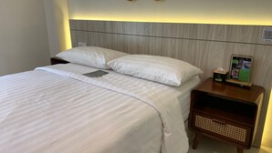 A neatly made bed with white linens, a wooden headboard, and a bedside table with a tissue box and a digital device. - The Baliem Hotel (Jimbaran)