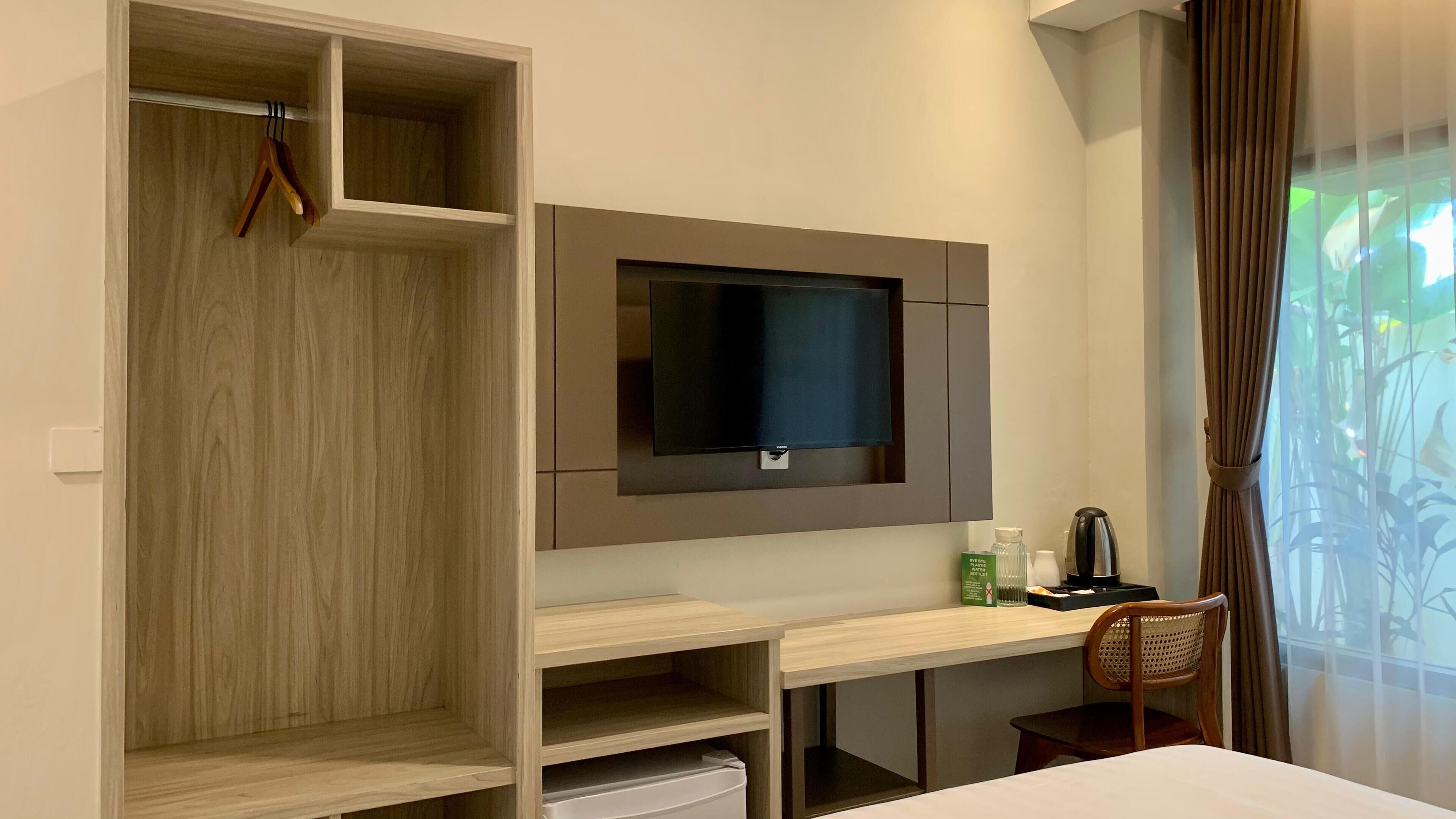A hotel room with a wooden wardrobe, a flat-screen TV mounted on the wall, a desk with a chair, and a window with curtains.
