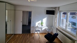 A modern room with a wooden floor, a black chair, a small table with a plant, a TV mounted on the wall, and a window with a view of buildings. - Vacation apartment on Lake Starnberg In the town center, just a few minutes from the lake (Seeshaupt)