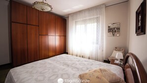 Une chambre avec une grande armoire en bois, un lit avec une literie à motifs, une table de chevet avec une lampe et un tableau encadré au mur.