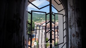 Une vue à travers une fenêtre ornée d’un cadre géométrique, dévoilant une rue bordée d’immeubles et un paysage vallonné en arrière-plan.