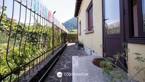 Un étroit sentier mène à un bâtiment dont le sol est en pierre, qui est entouré d’une clôture métallique ornée d’objets suspendus, et où l’on aperçoit une montagne en arrière-plan.