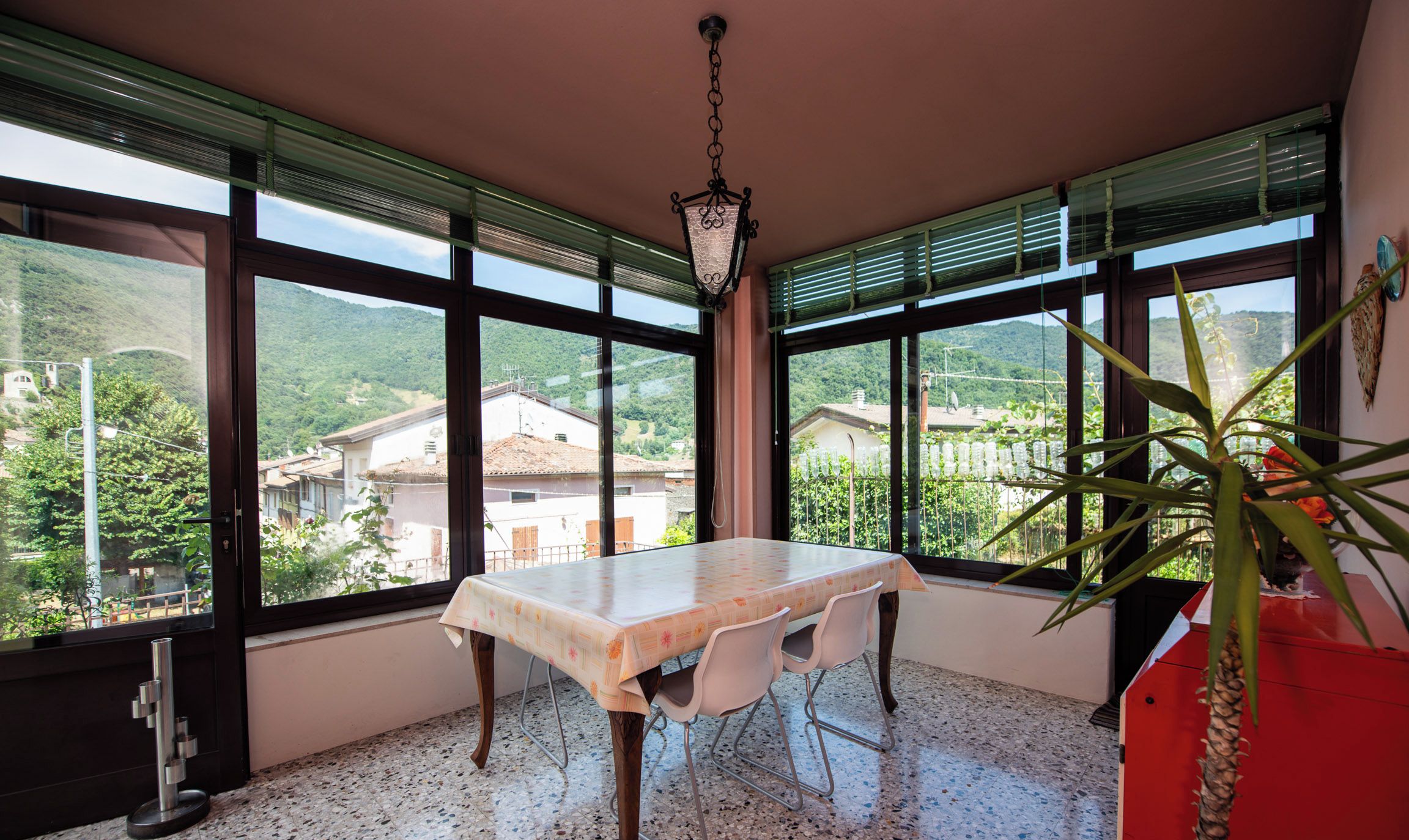 A dining area with a table and chairs, large windows with blinds, a hanging light fixture, and a potted plant.
