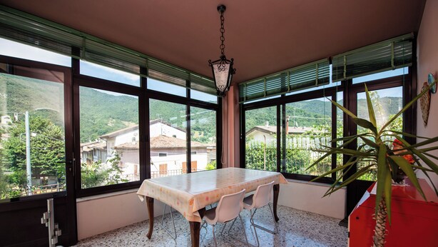 A dining area with a table and chairs, large windows with blinds, a hanging light fixture, and a potted plant.