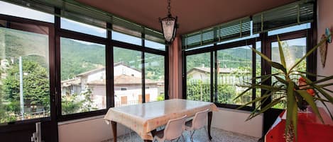 A dining area with a table and chairs, large windows with blinds, a hanging light fixture, and a potted plant.