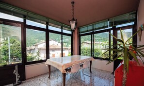 A dining area with a table and chairs, large windows with blinds, a hanging light fixture, and a potted plant. - Villa Fulvia (Vobarno)