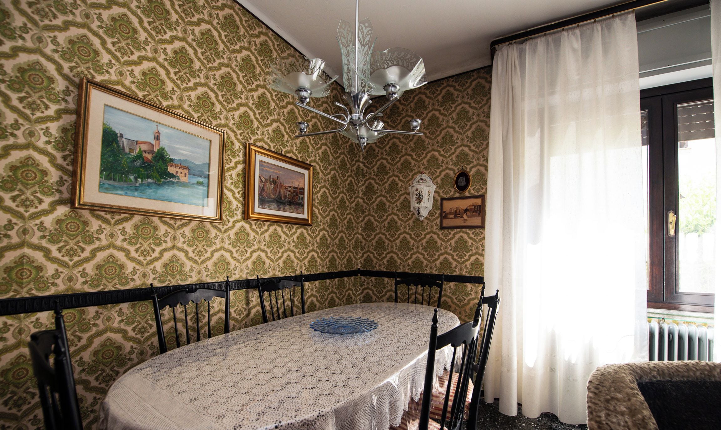 A dining area with a round table covered by a lace tablecloth, flanked by black chairs, against a patterned wallpaper backdrop.