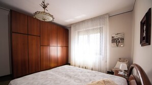 A bedroom with a large wooden wardrobe, a bed with patterned bedding, a nightstand with a lamp, and a framed picture on the wall.