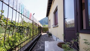 A narrow pathway leading to a building with a stone floor, a metal fence with hanging items, and a mountain in the background.