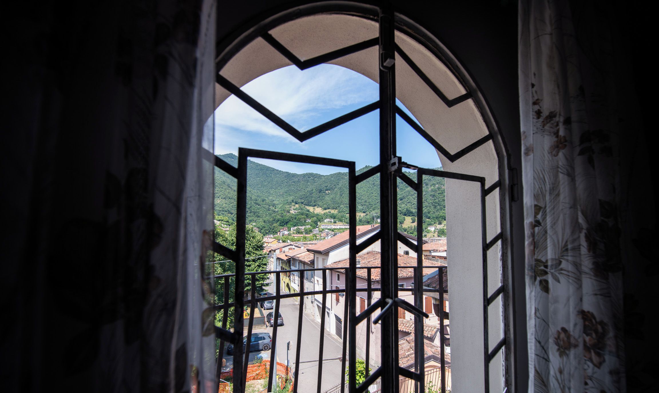 Una vista attraverso una finestra con cornice geometrica, che mostra una strada con edifici e un paesaggio collinare sullo sfondo.