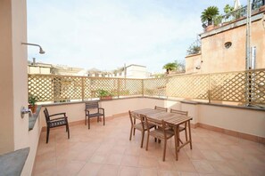 A rooftop terrace with a wooden table and chairs, a metal railing, and potted plants.