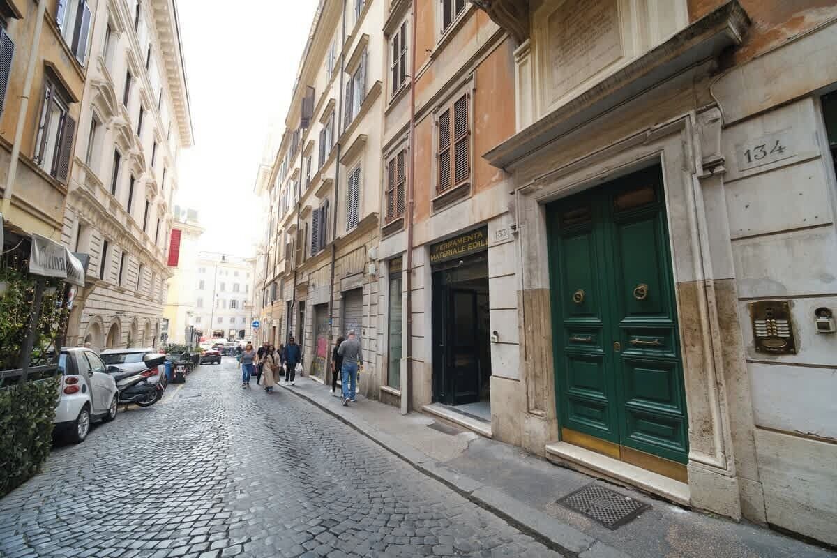 A cobblestone street with buildings, a green door, and a sign for 'FERRAMENTA MATERIALE ELLIOT.'