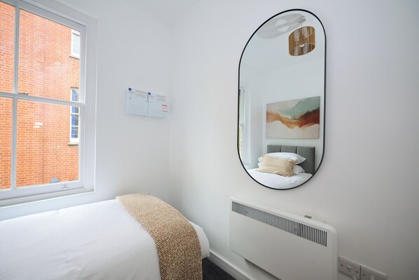 Une chambre d’hôtel avec un lit, un grand miroir, un radiateur et une fenêtre donnant sur un immeuble en briques.
