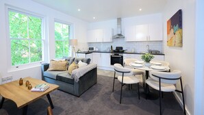 Eine moderne Küche mit weißen Schränken, ein Essbereich mit rundem Tisch und Stühlen sowie ein graues Sofa mit Kissen.