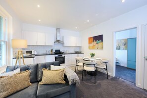 Una cocina moderna con armarios blancos, un comedor con mesa y sillas redondas, y un sofá gris con cojines decorativos.