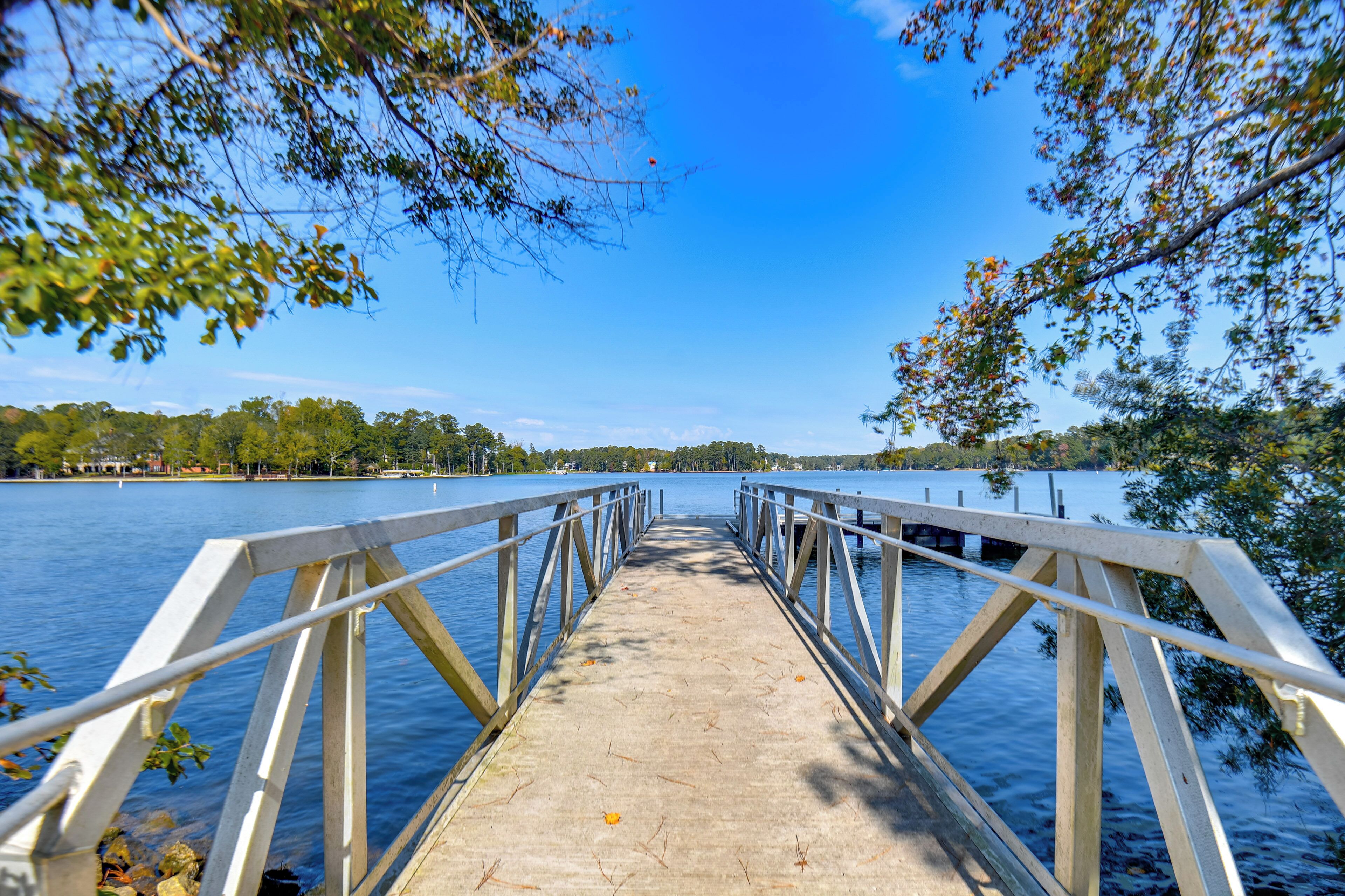 A concrete pier with metal railings extending over calm water, surrounded by trees and a clear sky.