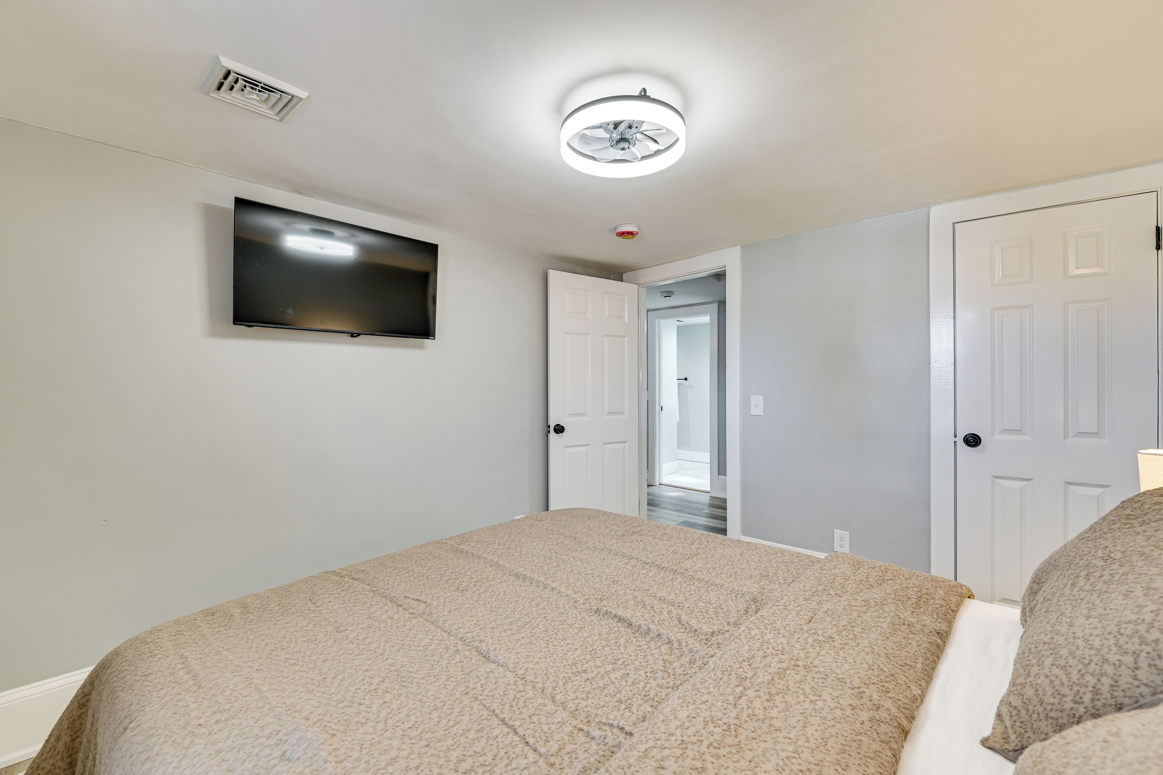 Un dormitorio con cama, ventilador de techo, televisor montado y una puerta que da a otra habitación.