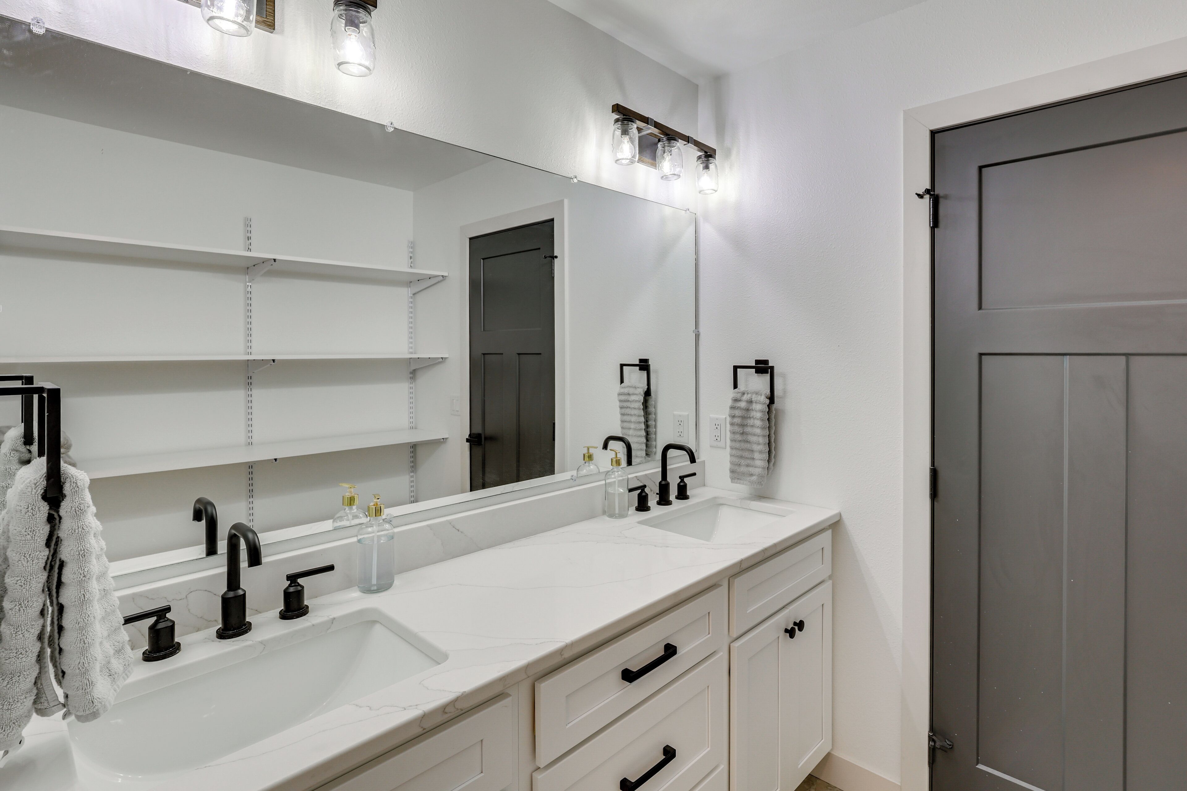 Une salle de bain avec un lavabo blanc, un robinet noir et un grand miroir. On y trouve des meubles blancs sous le lavabo et une serviette accrochée au mur.