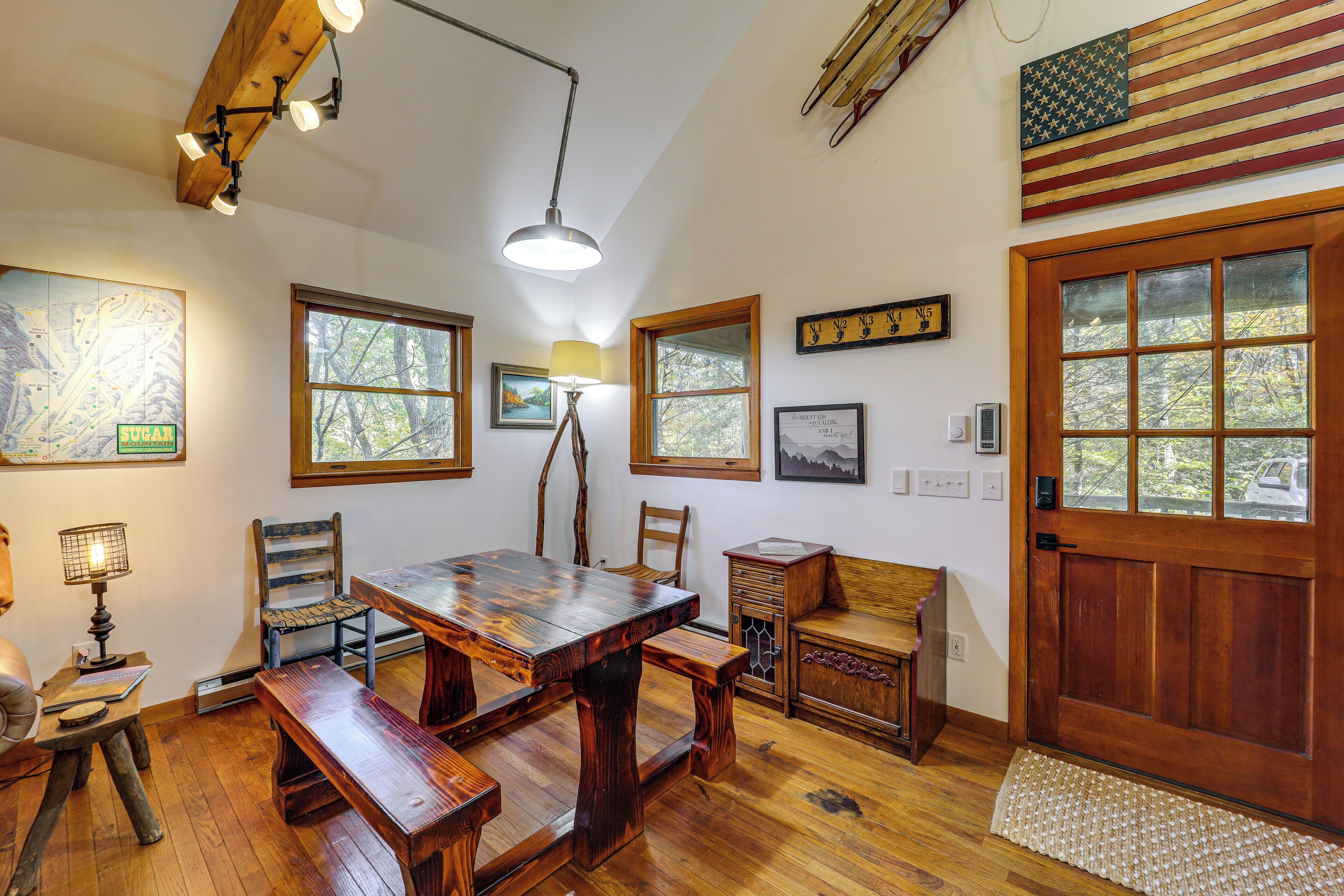 A wooden dining area with benches, a table, and a lamp. There are windows with views of trees and a door with a wooden panel.