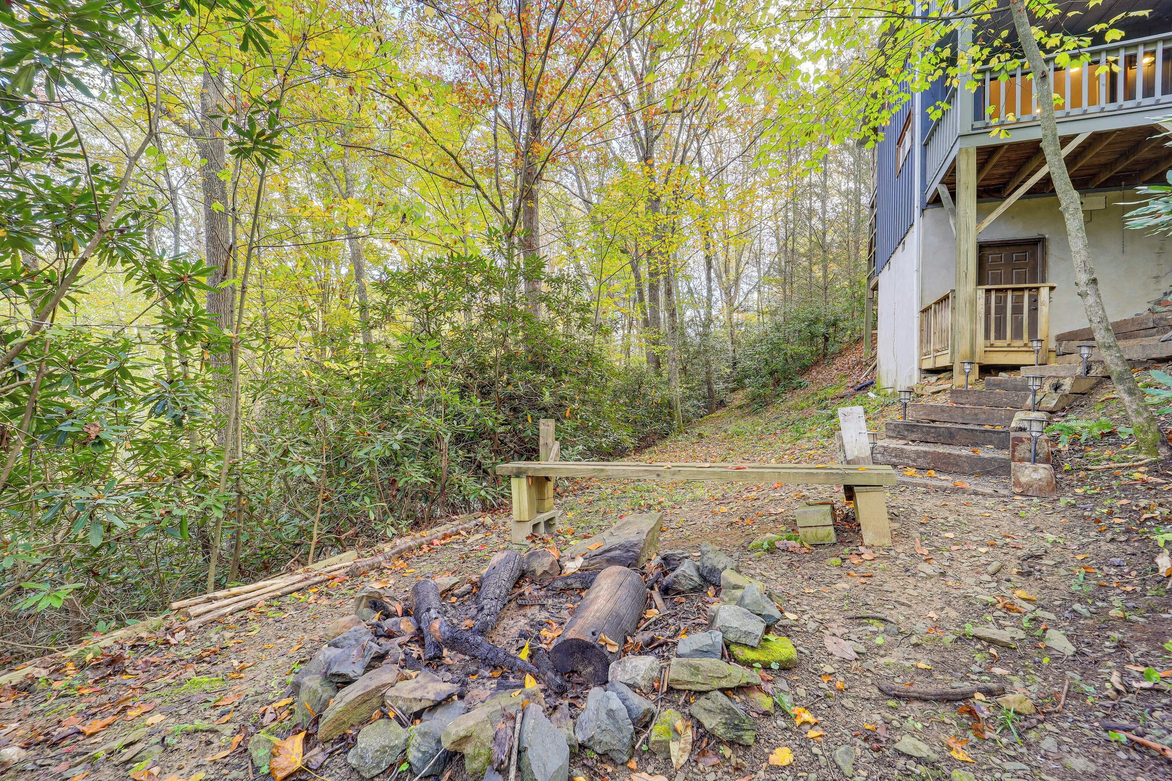 Eine Hütte mit einem Steinkamin, umgeben von Steinen und Baumstämmen, einer hölzernen Bank und einer Treppe, die zu einer Veranda mit Holzgeländer führt.