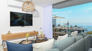 A modern living room with a large flat-screen TV, a wooden console table, and a sofa. A dining area with a table and chairs is visible through a glass partition, overlooking a scenic view. - vila Selavi (Tiberias)