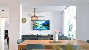 A modern living room with a large flat-screen TV mounted on the wall, a wooden dining table, and a grey sofa. - vila Selavi (Tiberias)