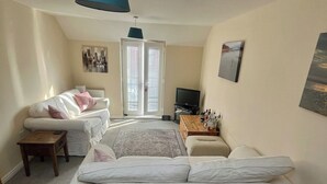 A cozy living room with a white sofa set, a small wooden table, a television, and a coffee table with bottles. - Entire Two Bedroom Detached Coach House (Exeter)