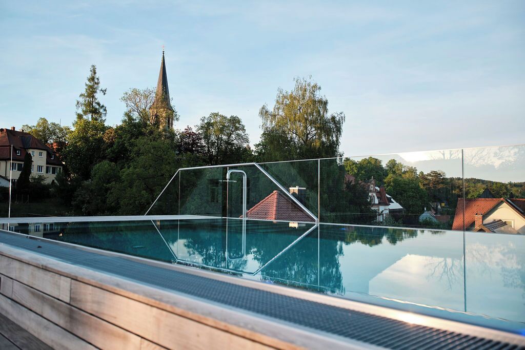 Ein Schwimmbad mit Glasbarriere, umgeben von Bäumen und Häusern, mit einem Kirchturm im Hintergrund.
