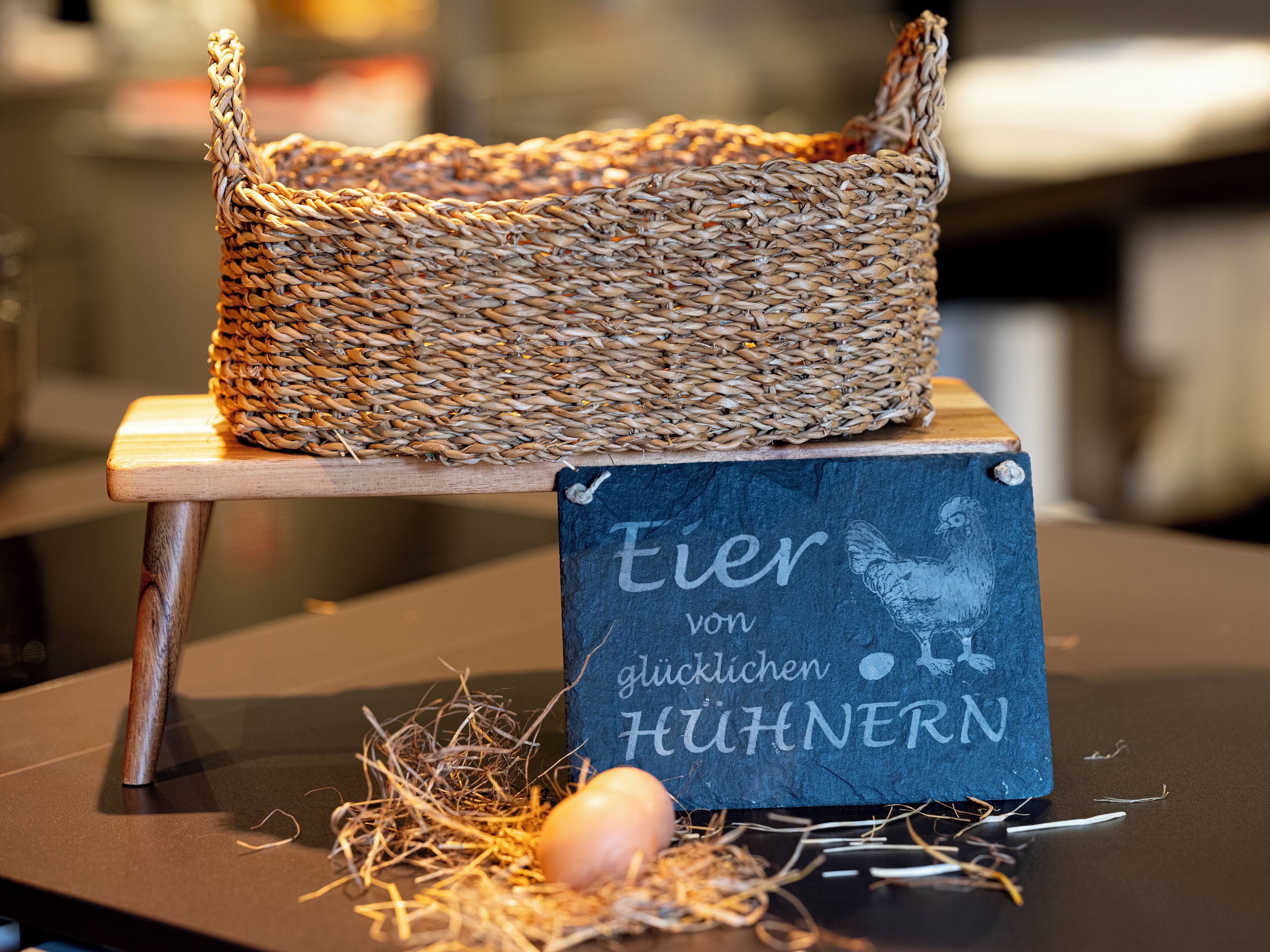 Un panier en osier posé sur une table en bois, avec une pancarte indiquant « Œufs de poules heureuses », et un nid contenant un œuf et de la paille.