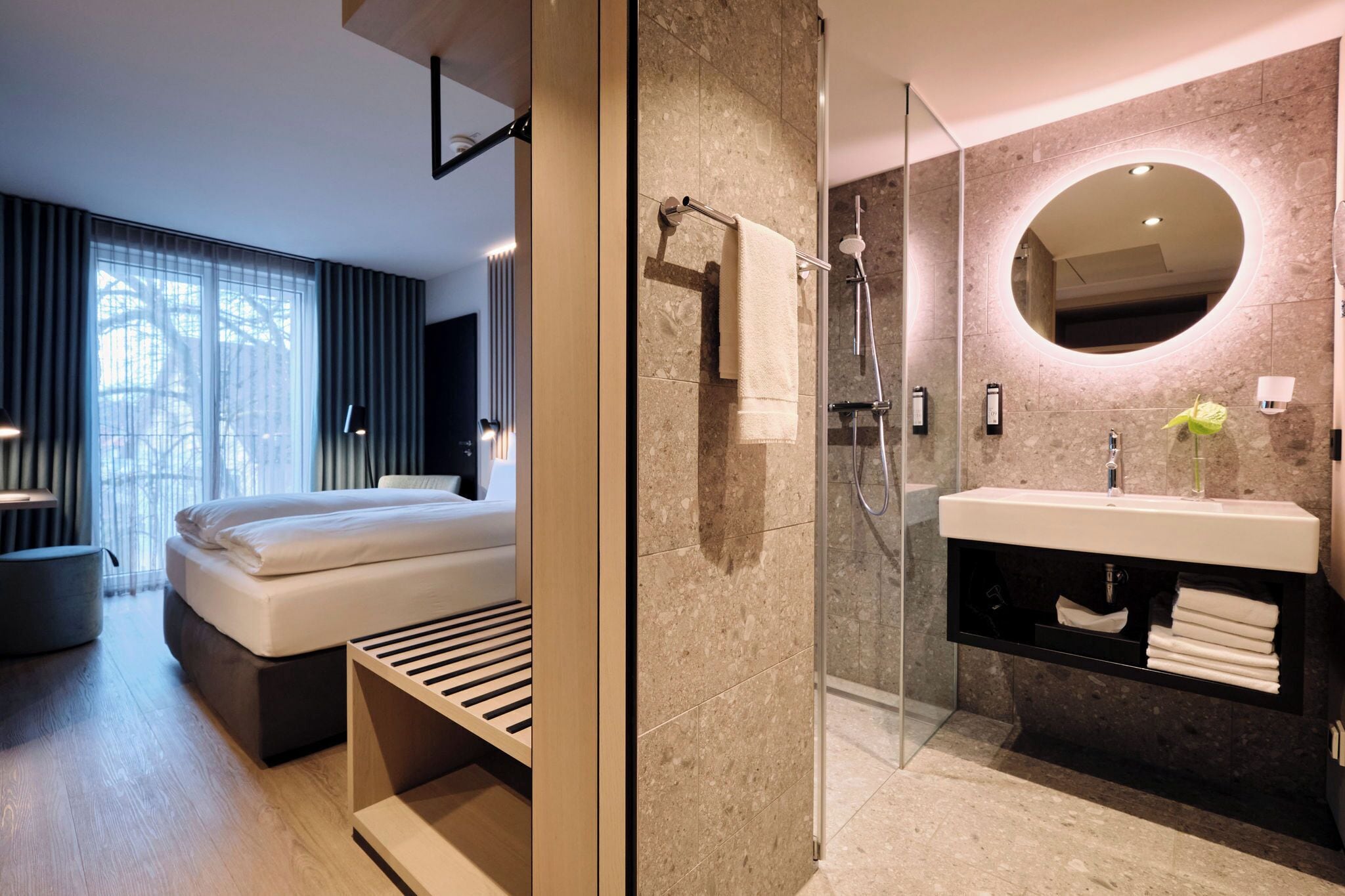 A modern bathroom with a glass-enclosed shower, a vanity with a mirror, and a bed in the adjacent room.