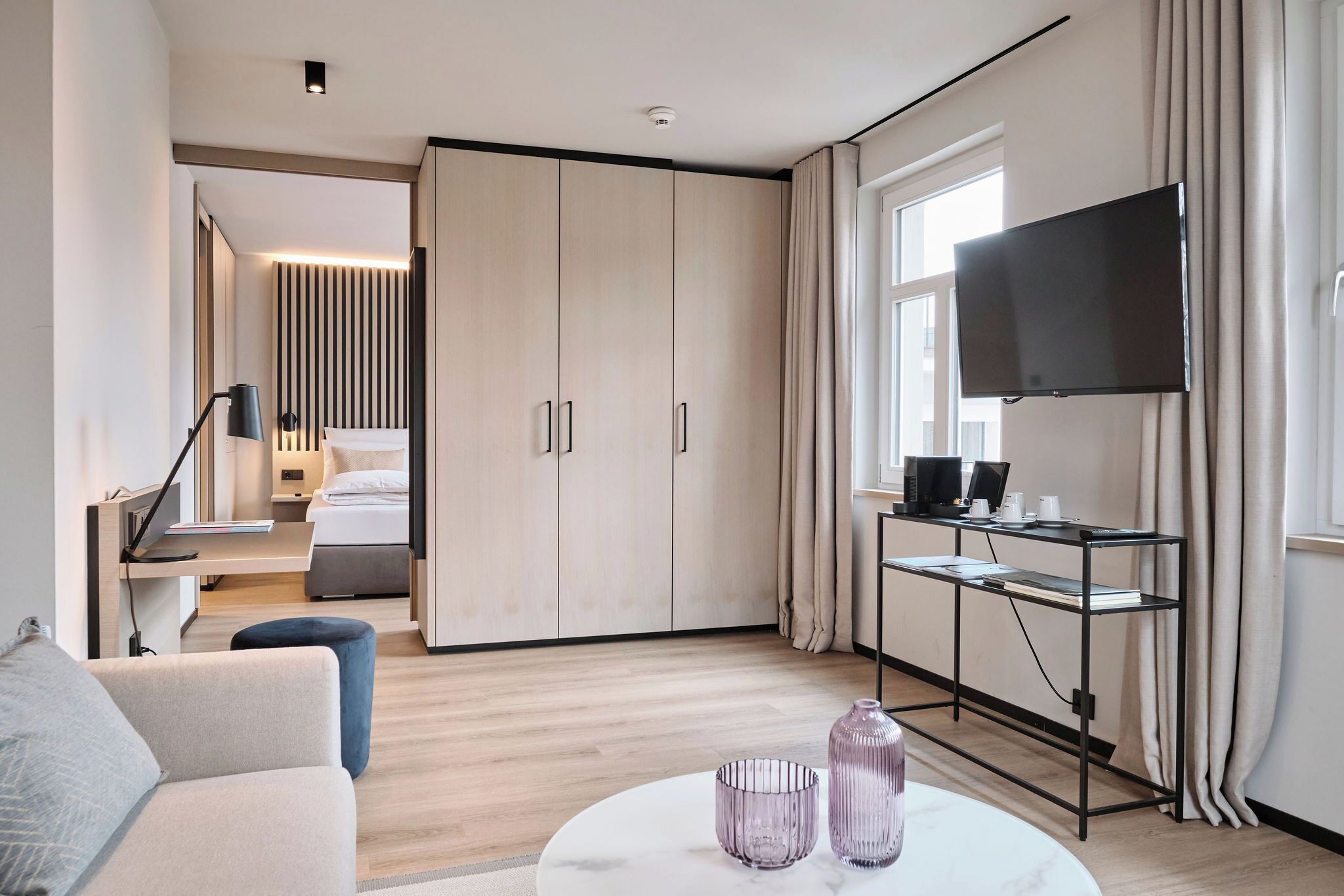 Una habitación de hotel moderna con cama, escritorio con computadora, televisor montado en la pared y un amplio armario.