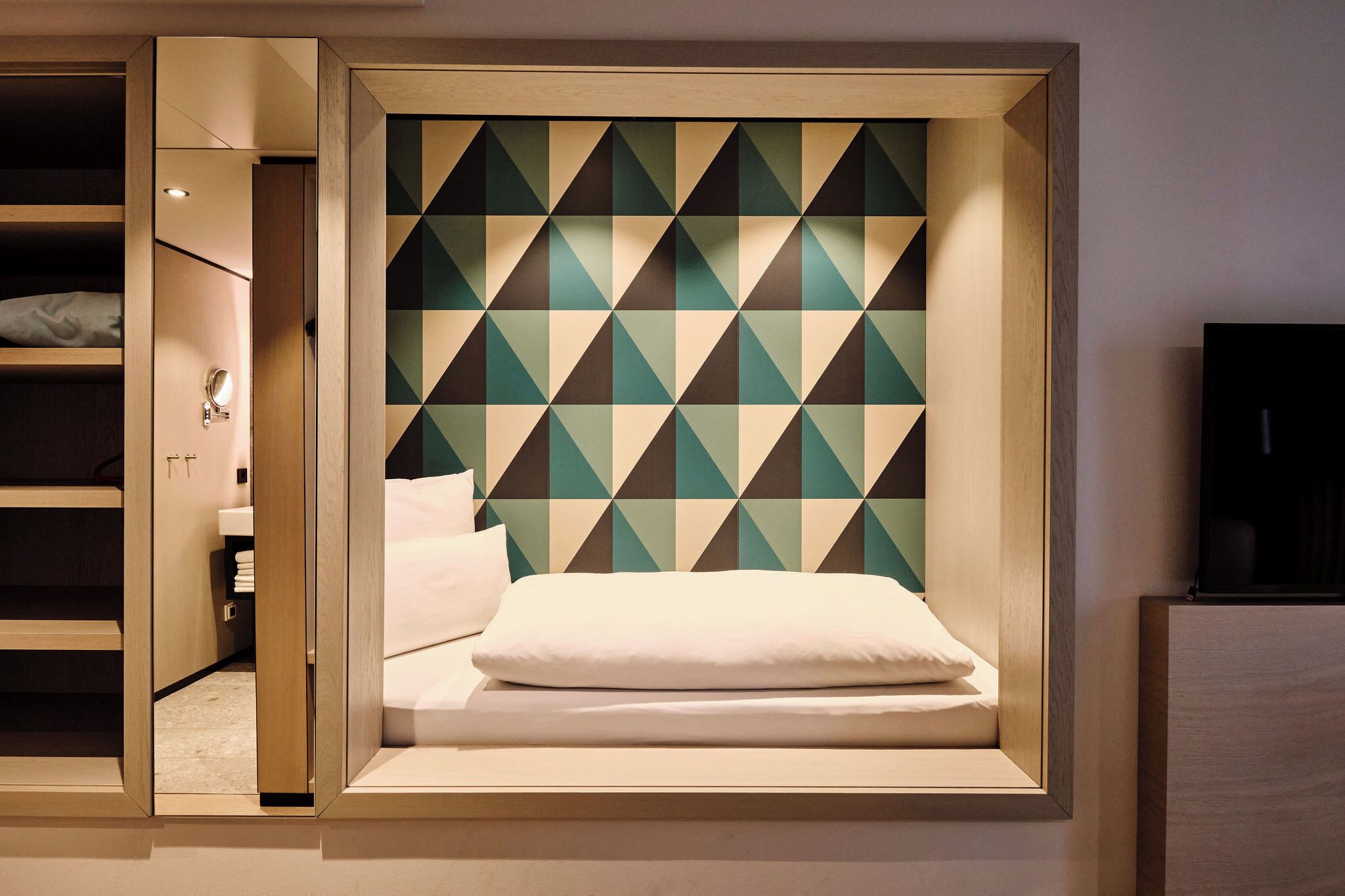 A modern room with a geometric patterned headboard, a bed with white bedding, and a visible bathroom with a mirror and towel rack.