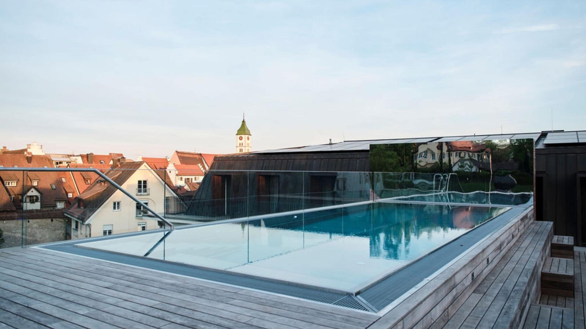 Une piscine sur le toit, protégée par une structure en verre, et entourée d’une terrasse en bois.