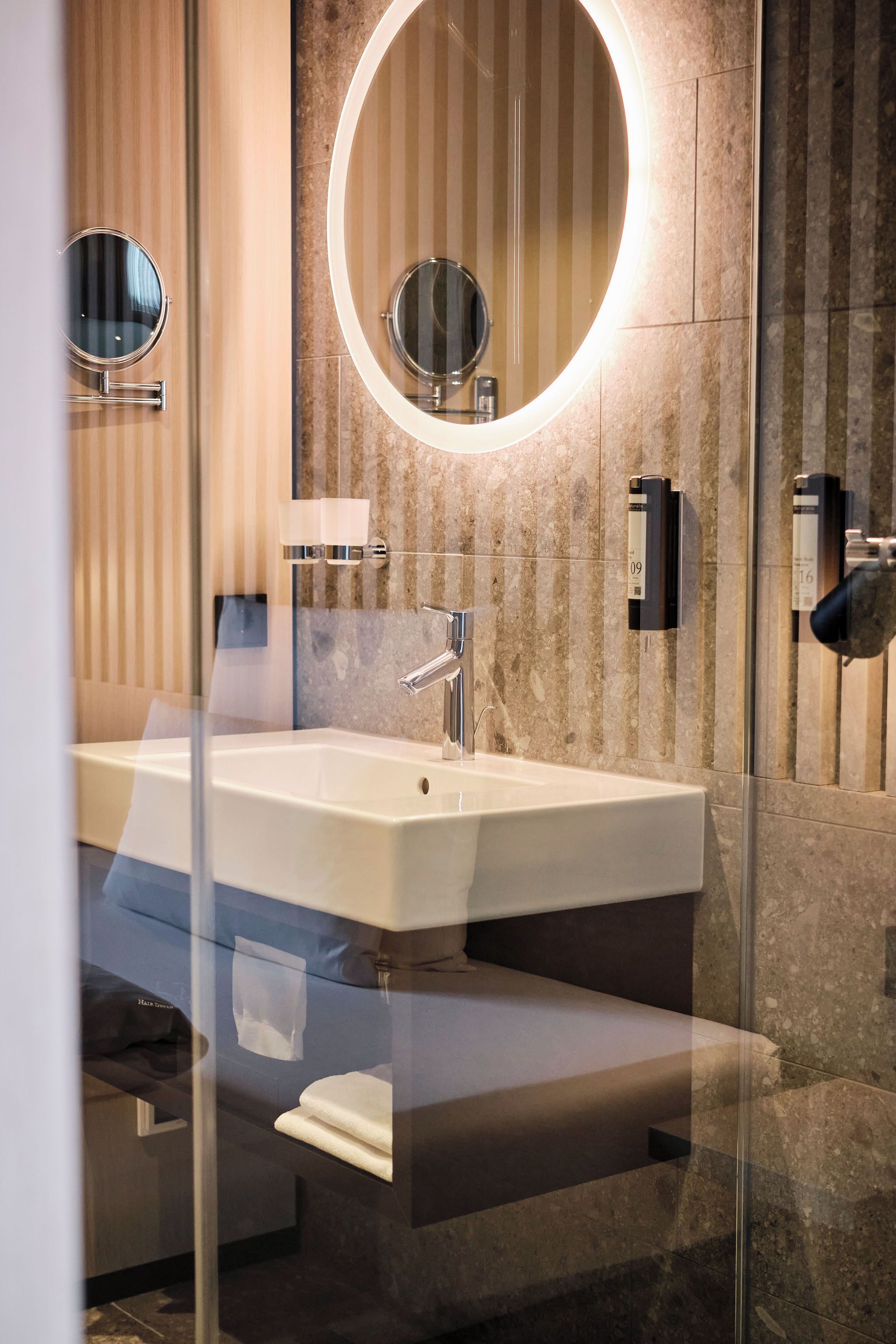A modern bathroom with a large rectangular sink, a round mirror with a light, and a glass shower enclosure.