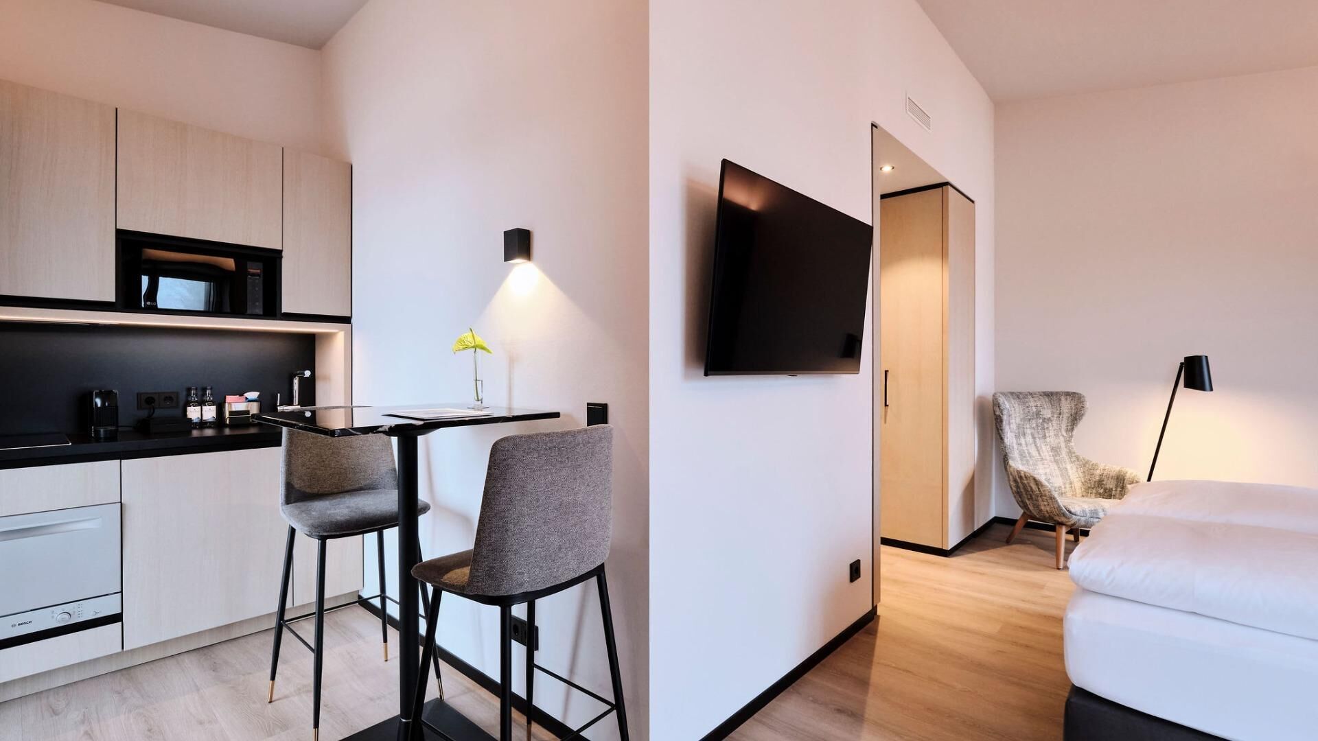 A modern hotel room with a kitchenette, a dining area, a TV mounted on the wall, and a seating area with a chair and a sofa.