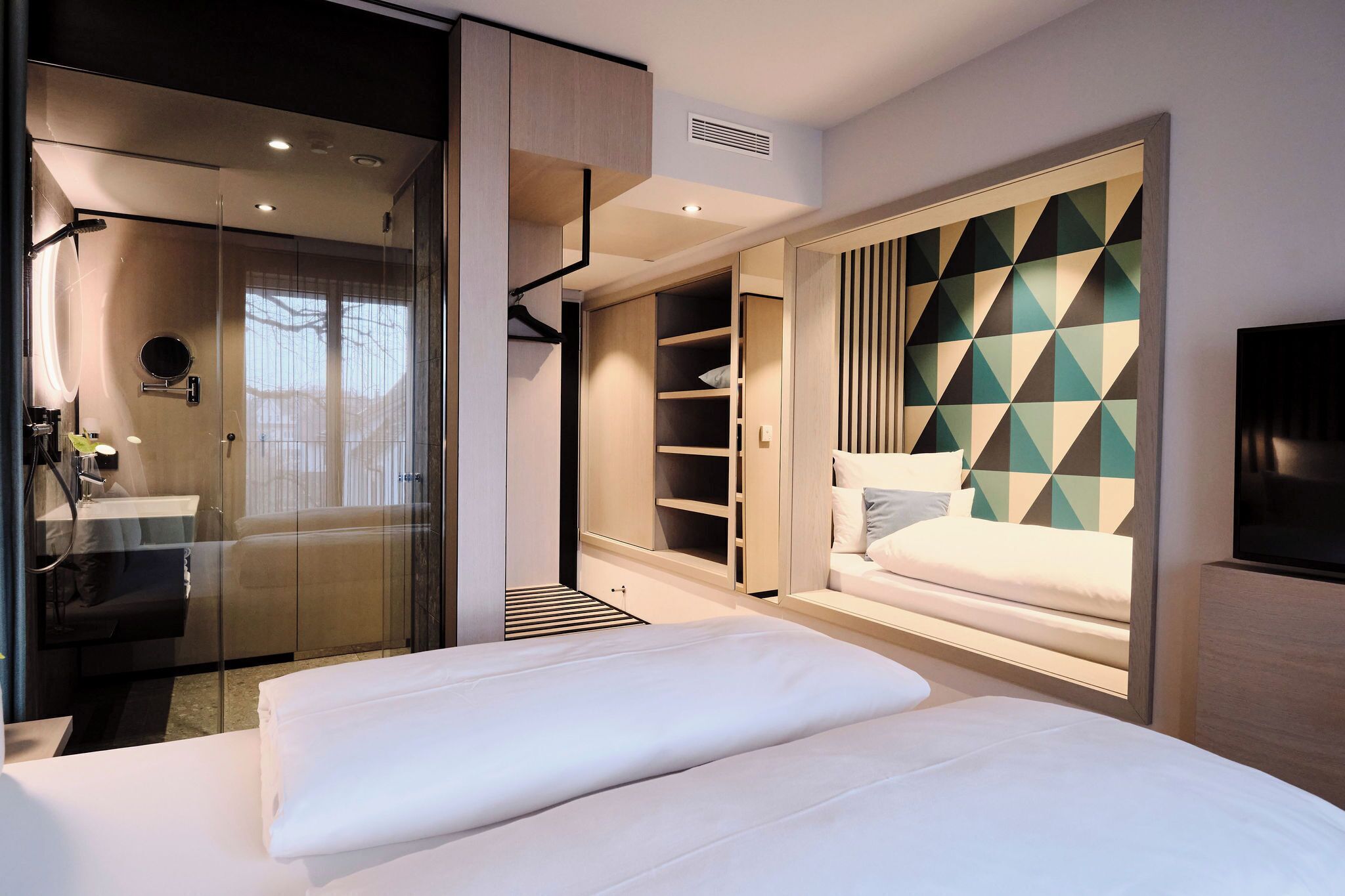 A modern hotel room with a large bed, a glass-enclosed shower area, a walk-in closet, and a TV on the wall.