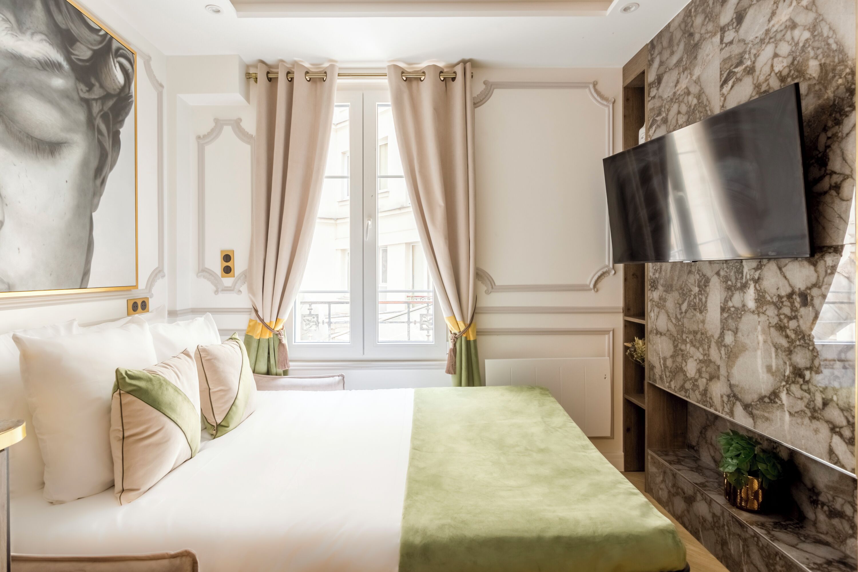 A hotel room with a bed, a flat-screen TV mounted on a marble wall, a window with curtains, and a wall-mounted shelf with a plant.