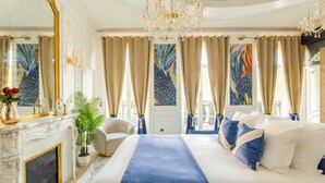 Une chambre à coucher luxueuse, dotée d’un grand lit, d’une cheminée, d’un lustre et d’œuvres d’art accrochées aux murs.