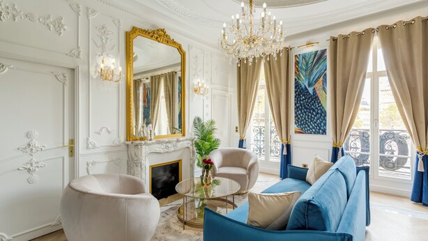Un salon luxueux aux murs blancs richement décorés, agrémenté d’un grand miroir, d’un lustre et d’un canapé bleu.