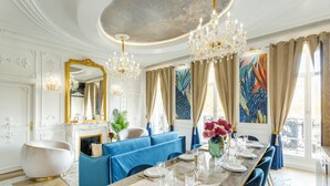 Une salle à manger avec une longue table dressée pour six personnes, des sièges recouverts d’un tissu bleu, des lustres et un grand miroir.